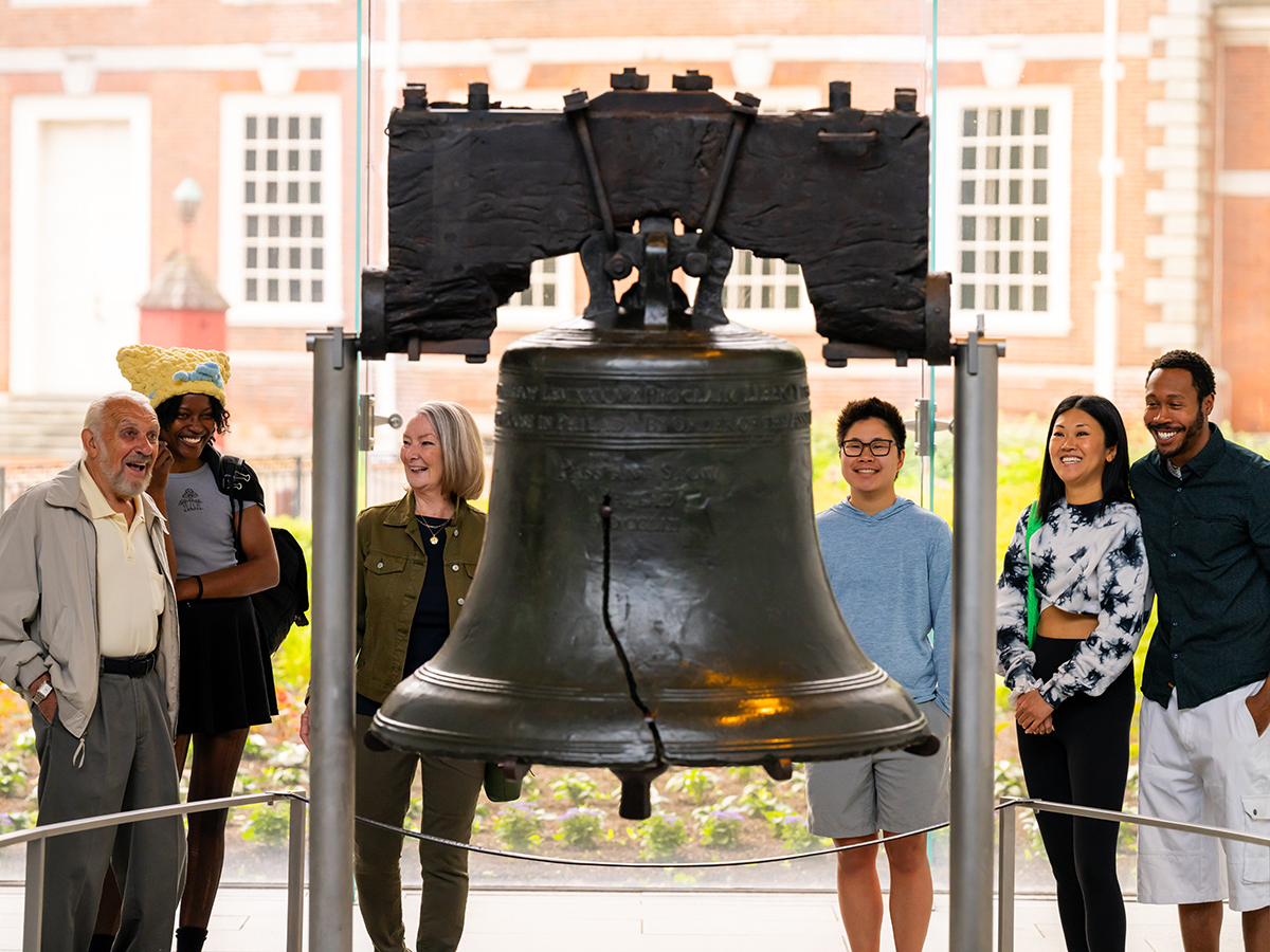 Visitors stand around and observe the Liberty Bell at Liberty Bell Center. Independence Hall is seen through the floor to ceiling windows behind the Liberty Bell.