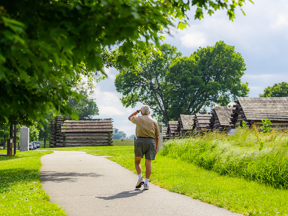 Man in korte broek loopt weg van de camera langs een geplaveid pad met groene bomen en historische hutten op Valley Forge National Historical Park.