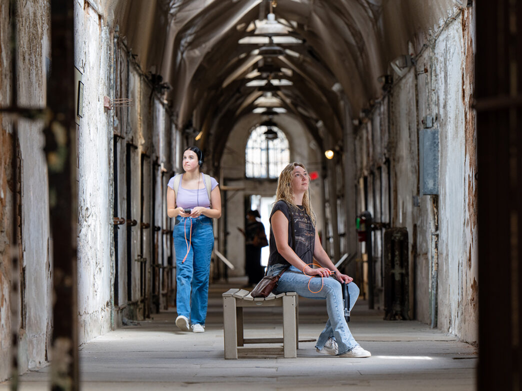 Duas pessoas, uma sentada em um banco e outra andando em um bloco de celas, usam fones de ouvido durante um tour de áudio no Eastern State Penitentiary.
