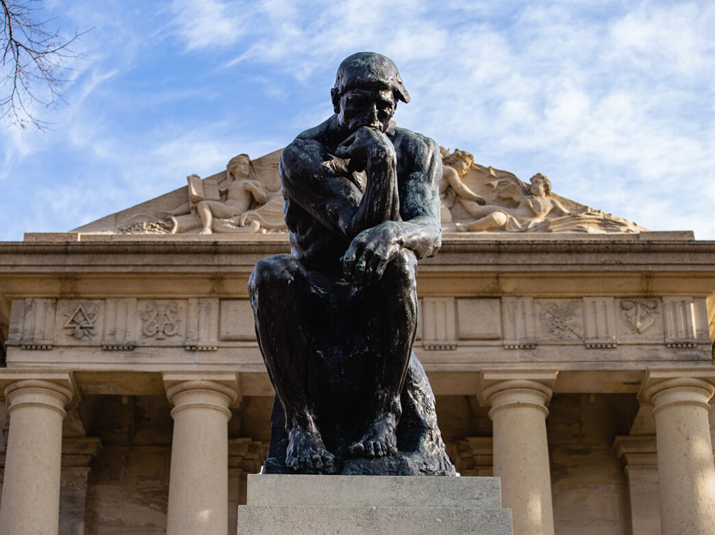 A close-up view of Rodin's "The Thinker" sculpture, a bronze figure seated in deep contemplation with his chin resting on his hand. The tan facade of the Rodin Museum can be seen in the background.