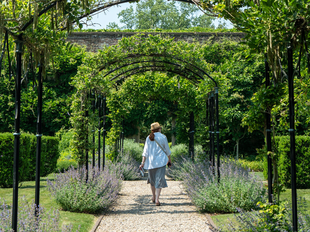 A person wearing a white shirt and a straw fedora hat walks away from the camera on a stone path. The path is surrounded by greenery.