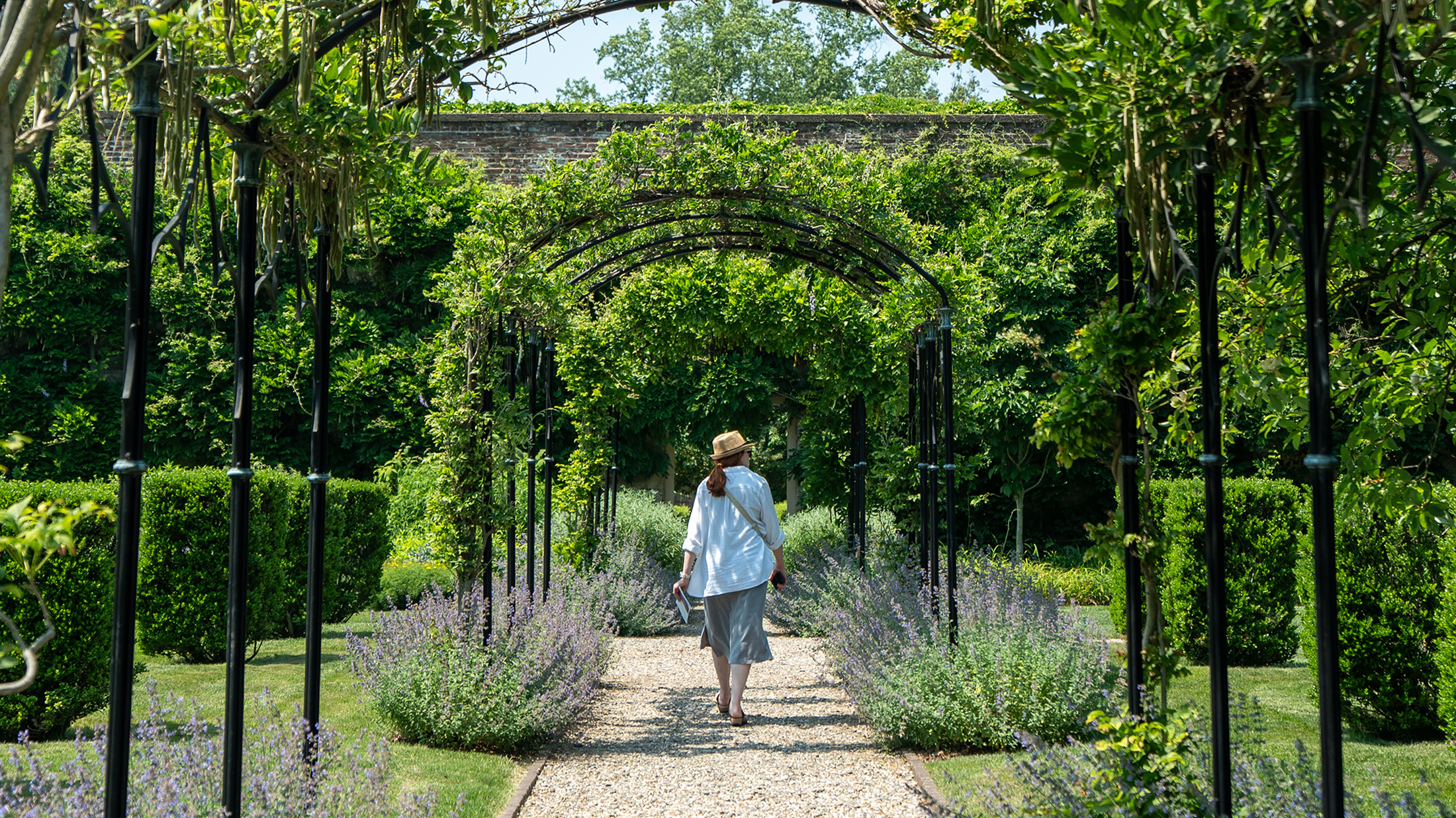 A person wearing a white shirt and a straw fedora hat walks away from the camera on a stone path. The path is surrounded by greenery.