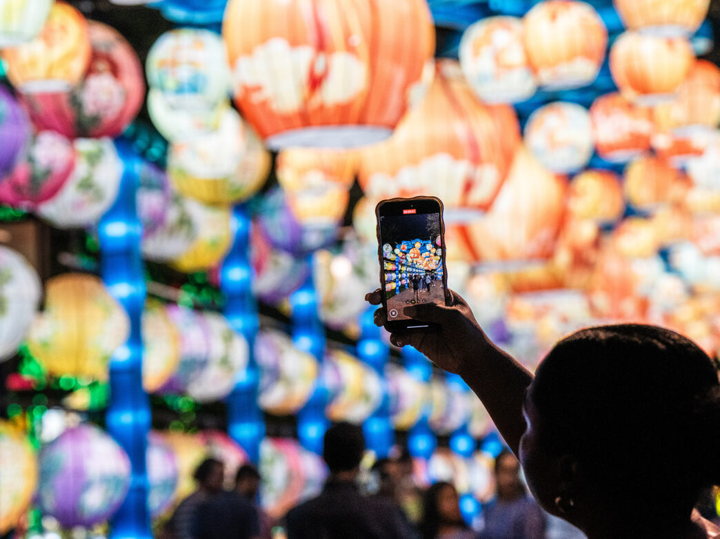 A person raises their phone to take a picture of the glowing, colorful lanterns suspended overhead in an tunnel at the Chinese Lantern Festival.