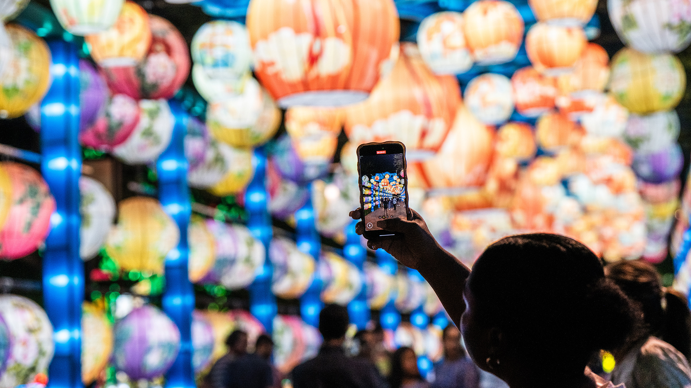A person raises their phone to take a picture of the glowing, colorful lanterns suspended overhead in an tunnel at the Chinese Lantern Festival.
