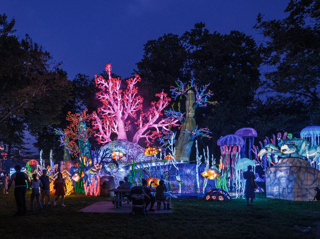 Crowds admire a massive light-up coral reef scene featuring glowing trees, colorful sea creatures and jellyfish lanterns against the night sky.