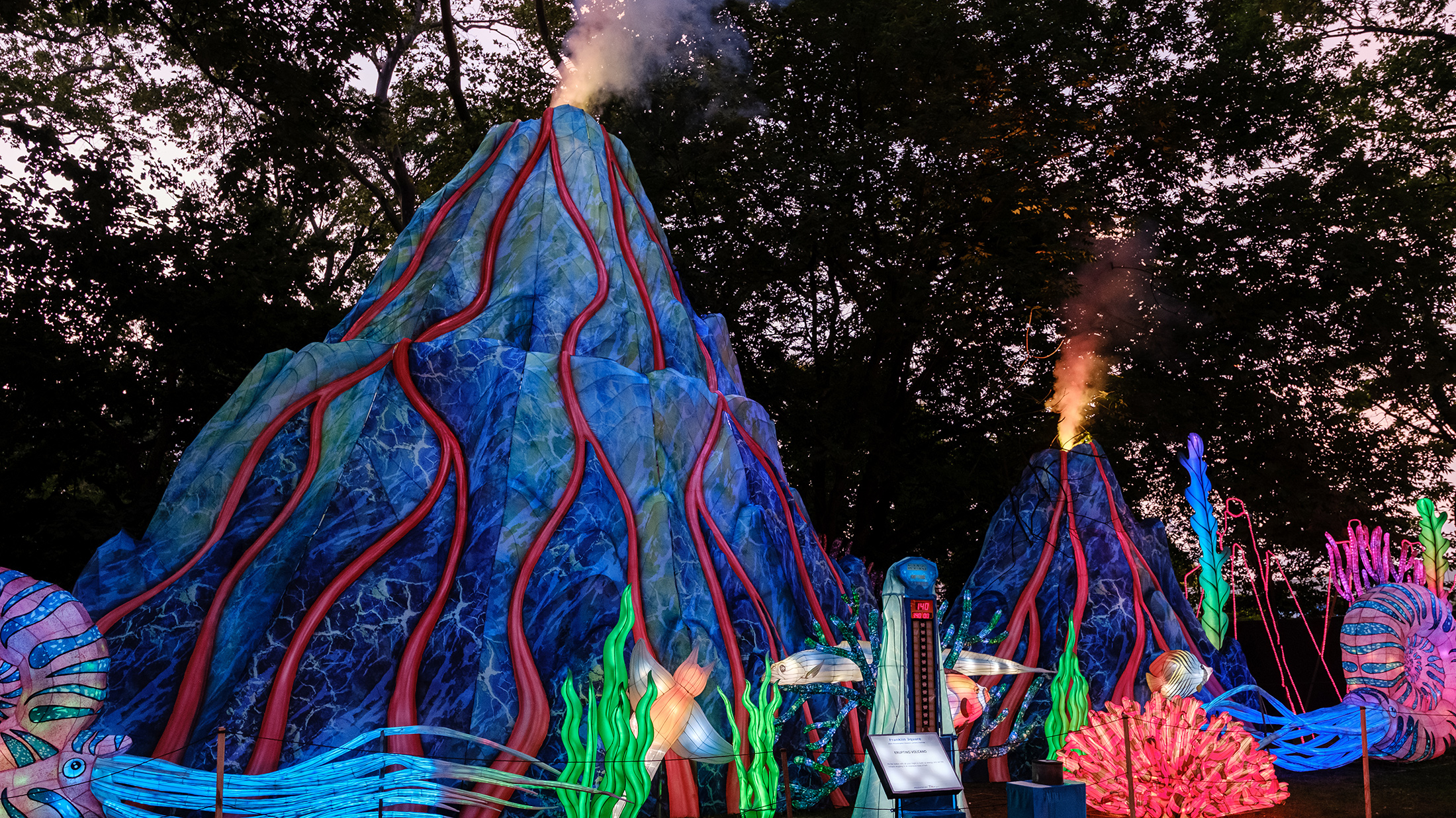 Towering, colorful volcano lanterns glow with blue and red hues as smoke puffs from the tops, surrounded by glowing sea creatures and coral lanterns.
