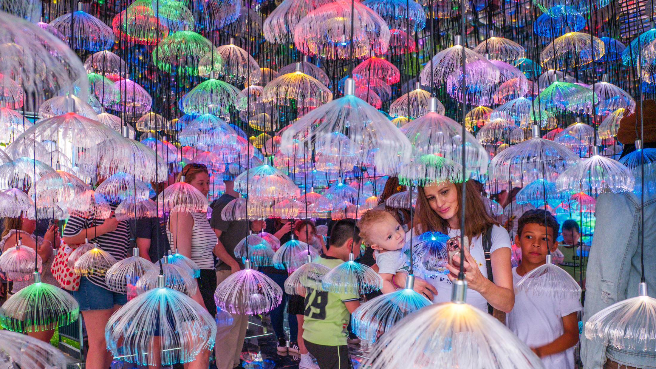 Visitors walk through a room filled with glowing multicolored fiber-optic jellyfish hanging from above.