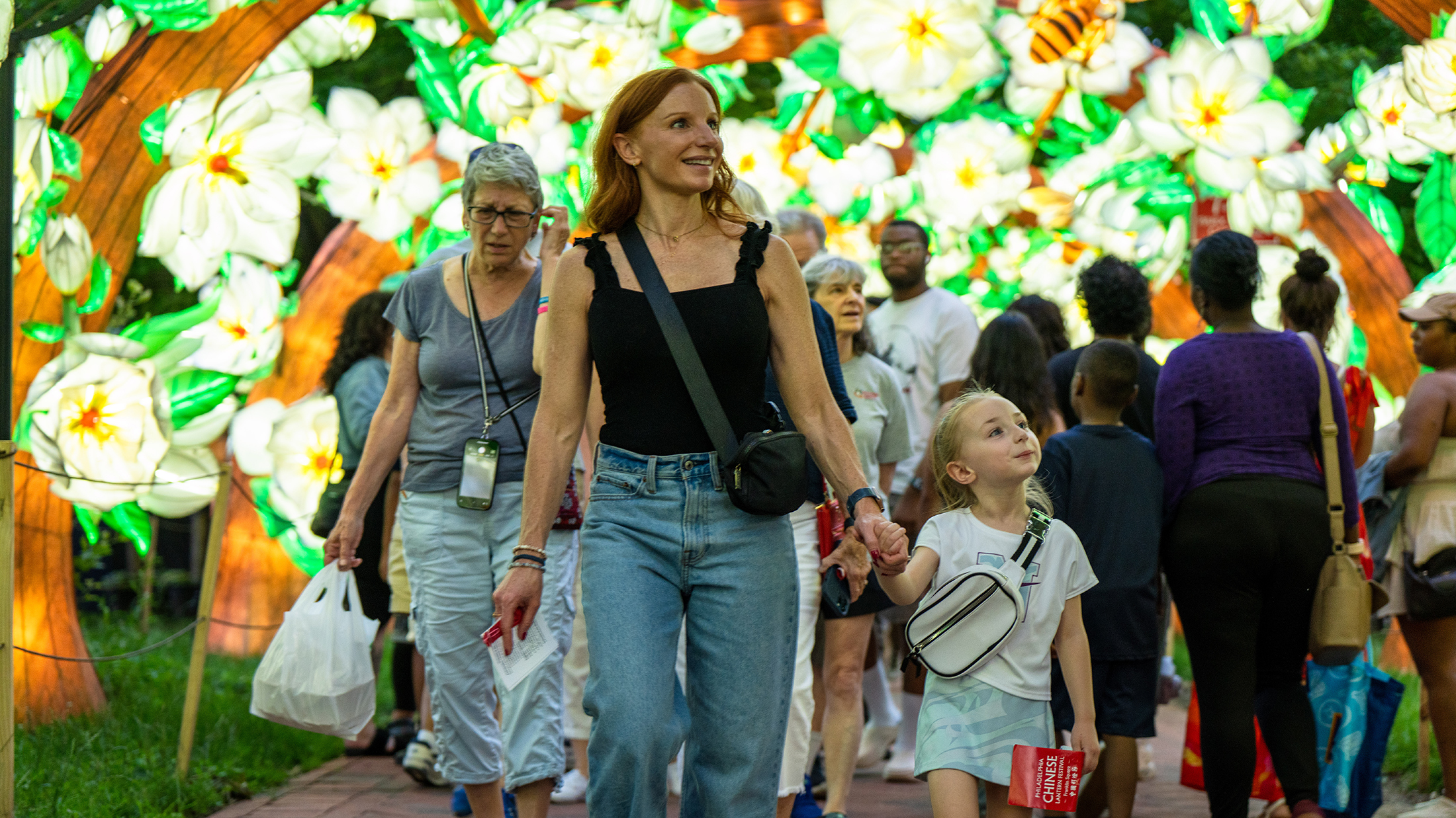 A woman and child walk hand-in-hand through a tunnel of glowing magnolia flower lanterns, surrounded by a crowd of people also enjoying the floral installation.