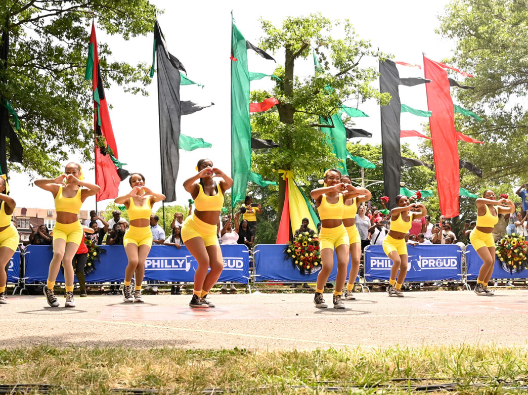 A coordinated group of dancers in bright yellow outfits perform in front of a crowd with red, green and black flags waving in the background.