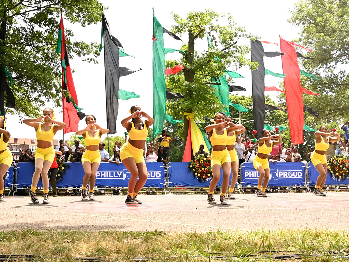 A coordinated group of dancers in bright yellow outfits perform in front of a crowd with red, green and black flags waving in the background.