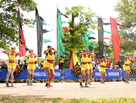 A coordinated group of dancers in bright yellow outfits perform in front of a crowd with red, green and black flags waving in the background.