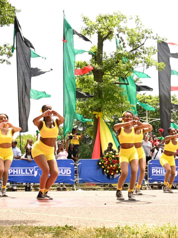 A coordinated group of dancers in bright yellow outfits perform in front of a crowd with red, green and black flags waving in the background.