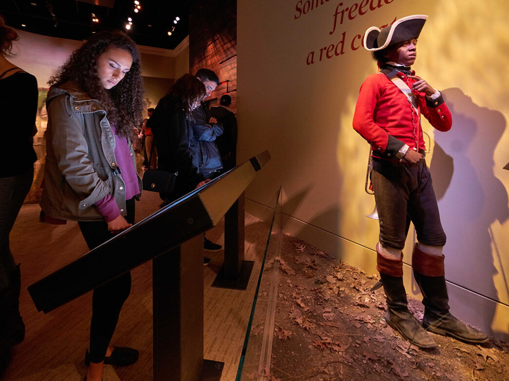 A woman uses a touchscreen at a museum exhibit beside a life-sized mannequin dressed as a soldier in a redcoat uniform.
