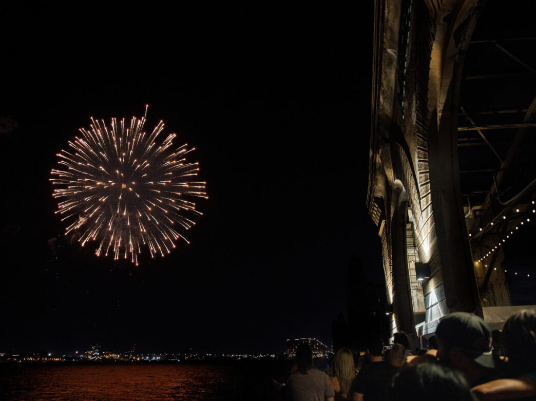A golden firework explodes over the Delaware River as people gather under the industrial overhang of Cherry Street Pier at night.