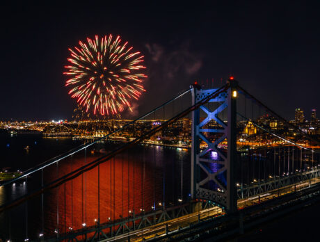 A bright burst of fireworks illuminates the dark sky above the Benjamin Franklin Bridge with Philadelphia's skyline and Delaware River glowing beneath it.
