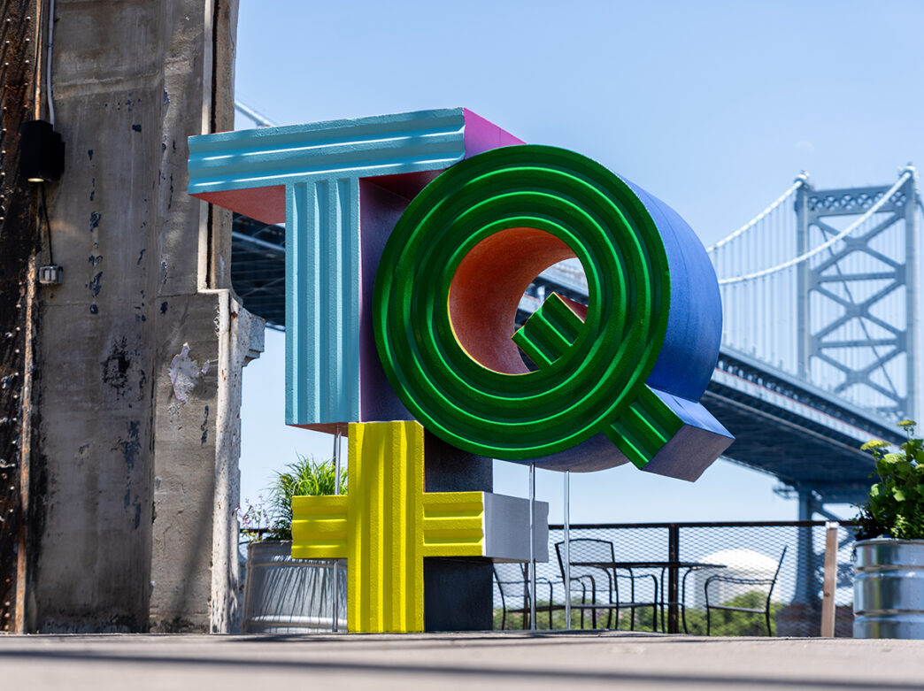 A multi-colored sculpture that says "TQ+" stands outdoors. The Benjamin Franklin Bridge is visible in the background.