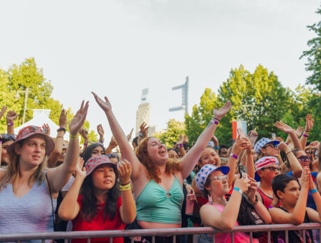 A lively crowd of people stand behind a metal railing, cheer and raise their hands in excitement while attending the July 4th concert in Philadelphia.