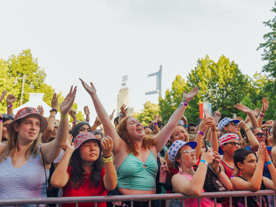A lively crowd of people stand behind a metal railing, cheer and raise their hands in excitement while attending the July 4th concert in Philadelphia.
