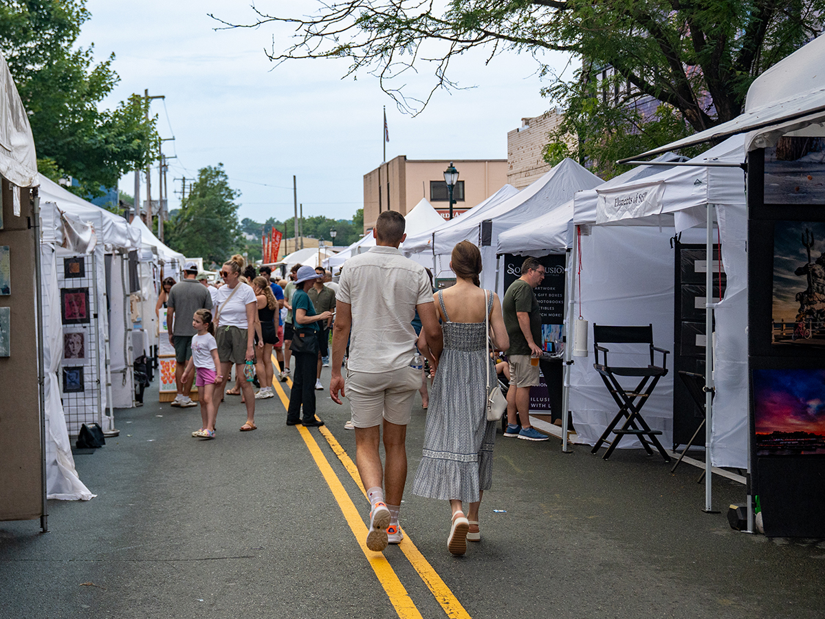 A couple walks hand-in-hand down a street lined with white vendor tents at the bustling Manayunk Arts Festival.