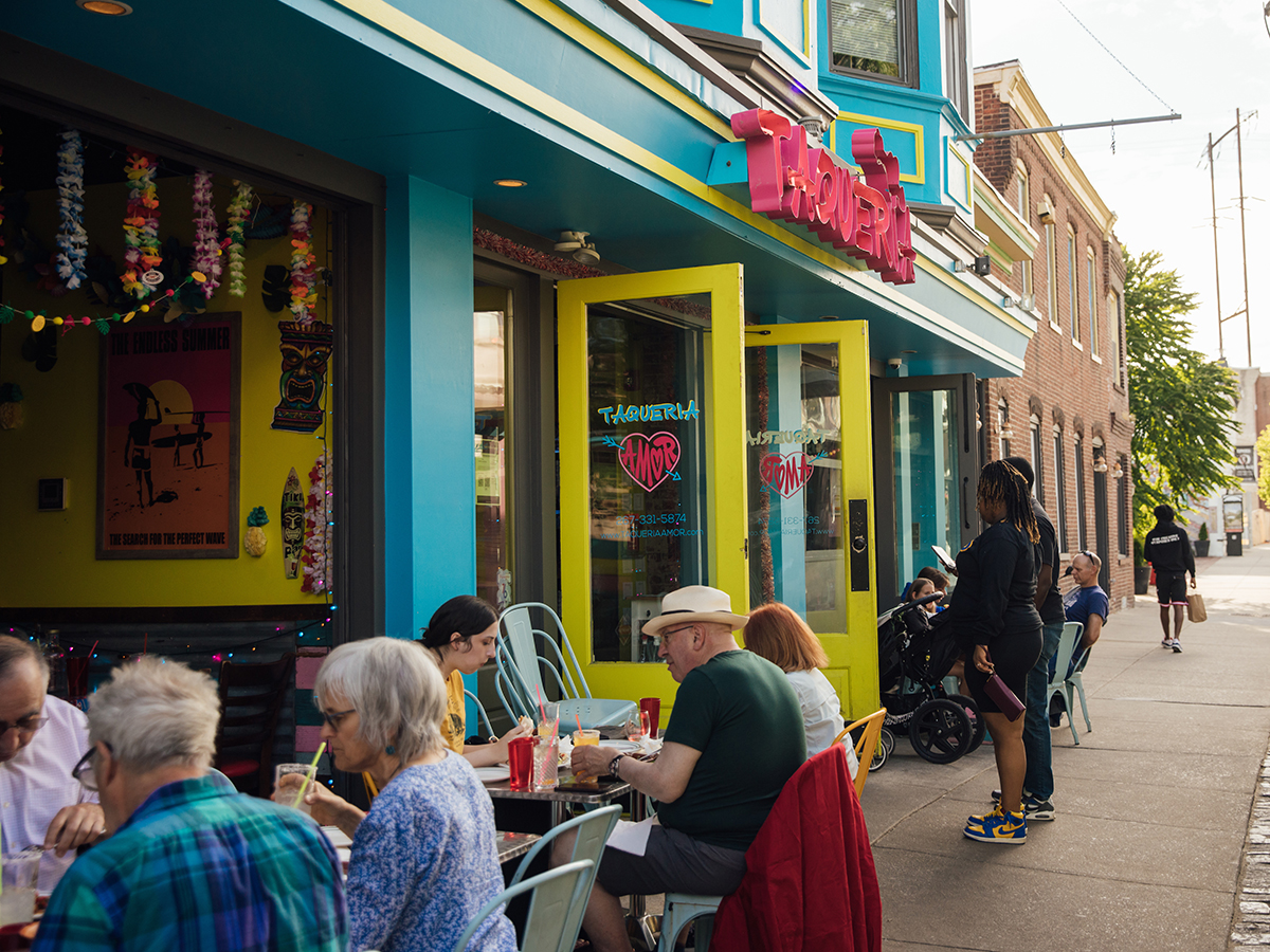 Diners enjoy food and drinks at colorful outdoor tables in front of the bright blue and yellow facade of Taqueria Amor in Manayunk.