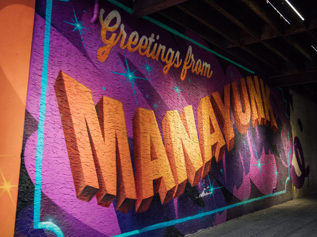 Une fresque colorée sous un pont affiche en lettres orange vives sur fond violet : " Salutations de Manayunk ".