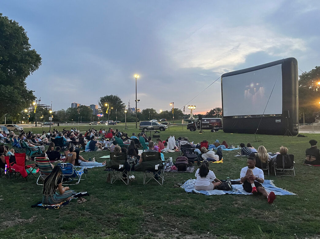 People lounge on blankets and lawn chairs in front of a large inflatable movie screen at The Oval, watching an outdoor film as the sun sets.