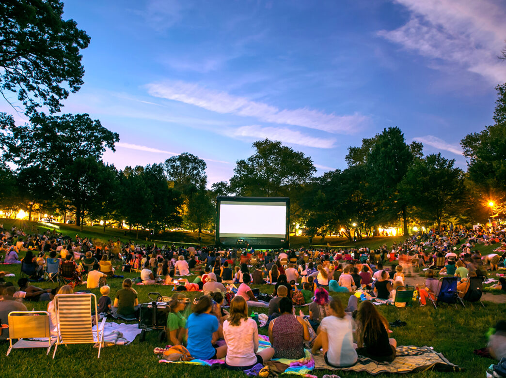 A large crowd gathers on blankets and lawn chairs to watch a movie on a giant screen under the evening sky at Clark Park.