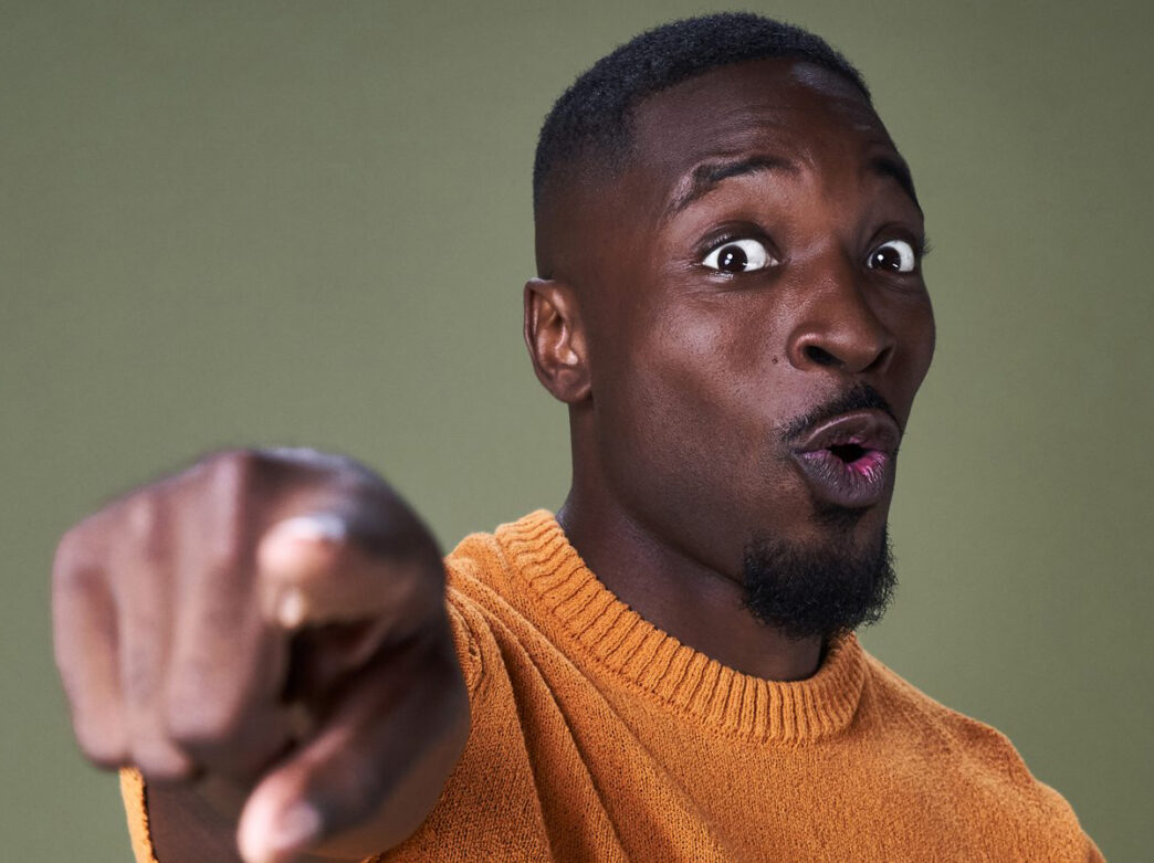 Comedian Preacher Lawson wears an orange sweater and poses for the camera, pointing at the camera with wide-eyes.
