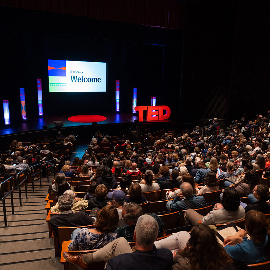 A packed theater audience sits facing a brightly lit stage, where a screen reads "Welcome" alongside TED Democracy branding and colorful pillars.