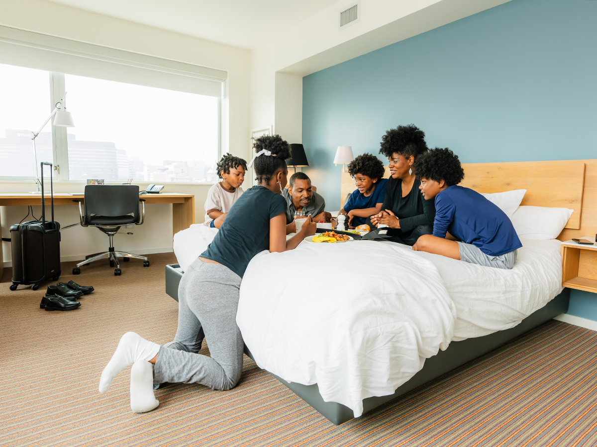 A smiling family of six sits together on a hotel bed enjoying breakfast.