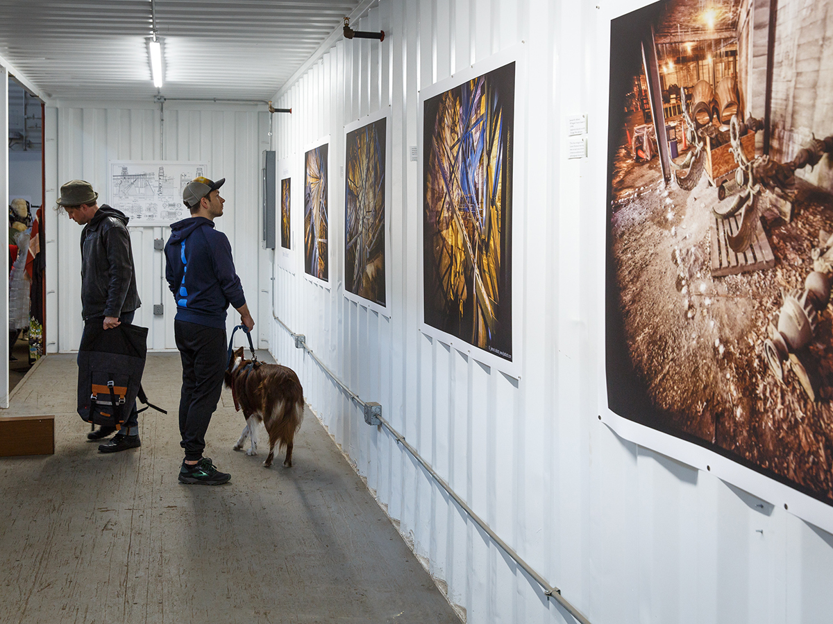 Two people, one with a dog on a leash, admire large scale photographs and artwork displayed on white walls of a shipping container gallery at Cherry Street Pier.