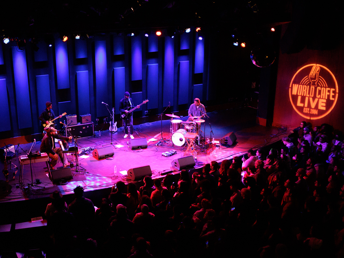 A band performs on a stage with pink and blue lighting while a packed audience watches from below.