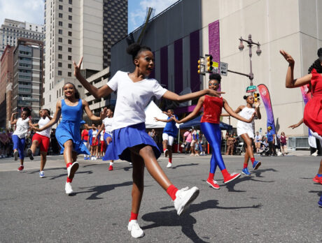 A group of young dancers in red, white and blue outfits perform a dance routine in the middle of Broad Street during the Avenue of the Arts Block party. Philadelphia's City Hall is visible in the background.