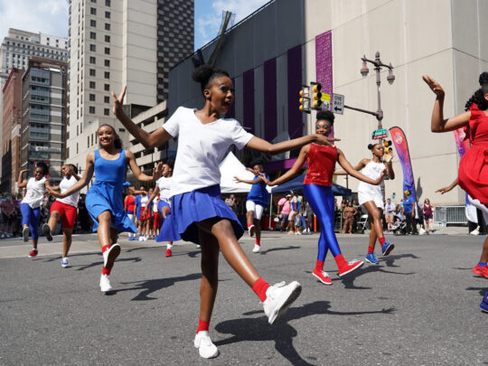 Um grupo de jovens dançarinos vestidos com roupas vermelhas, brancas e azuis executa uma coreografia no meio da Broad Street durante a festa Avenue of the Arts Block. O City Hall da Filadélfia é visível ao fundo.