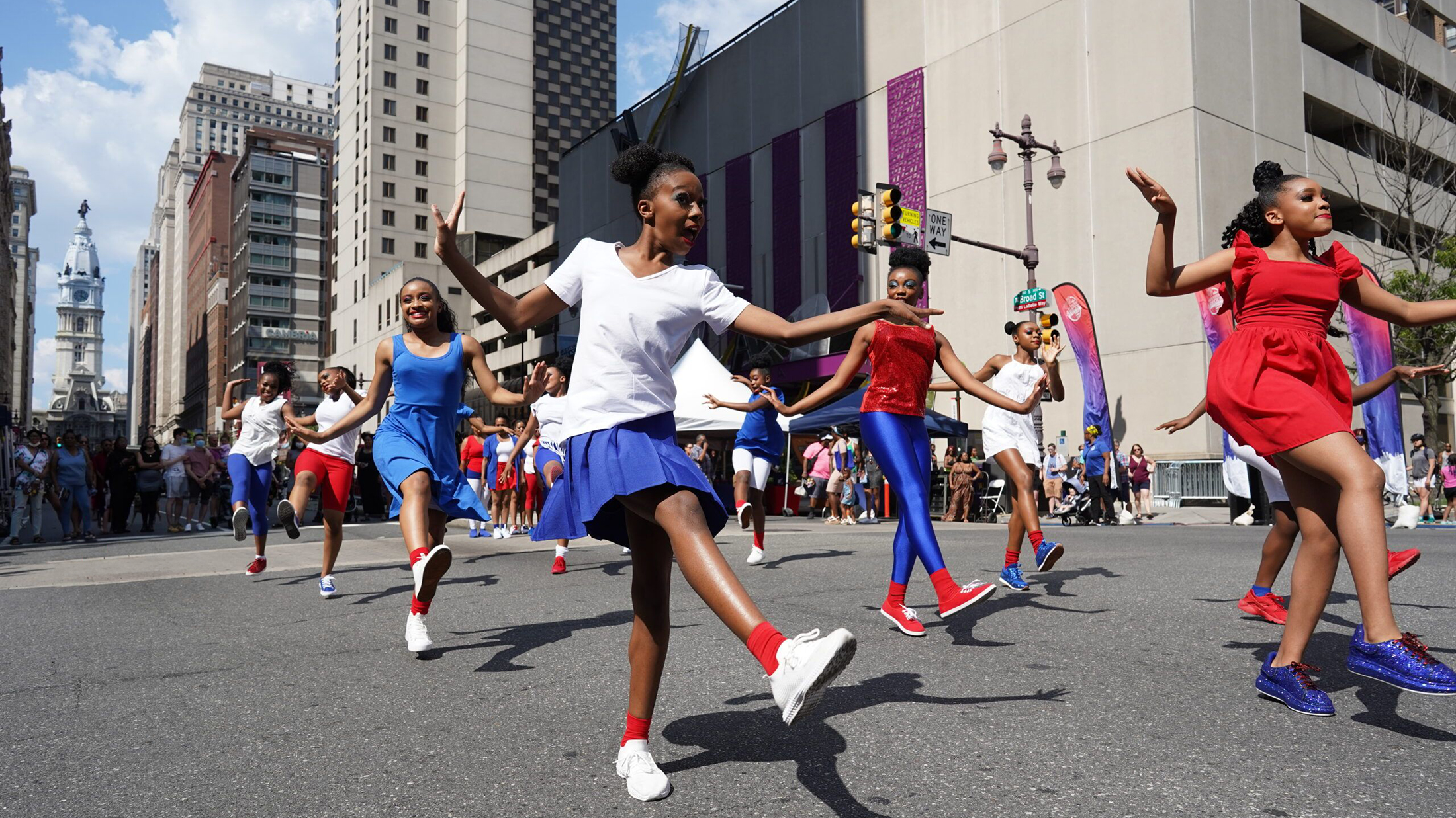 Un groupe de jeunes danseurs vêtus de costumes rouges, blancs et bleus exécutent une chorégraphie au milieu de Broad Street pendant la fête de quartier Avenue of the Arts Block. Le City Hall de Philadelphie est visible à l'arrière-plan.