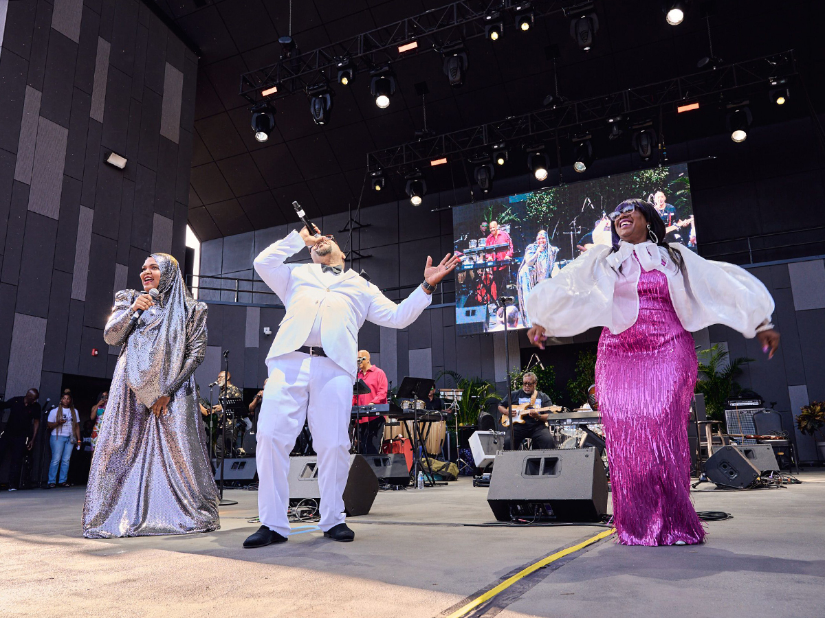 Three vocalists in shimmering silver, white and pink outfits sing and dance on stage backed by a full band.
