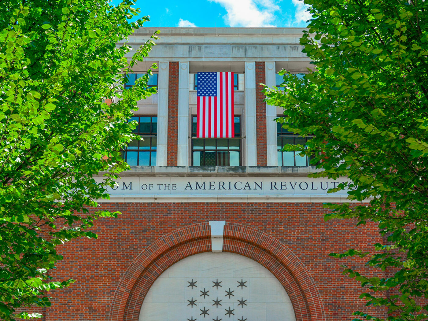 A large American flag hands above the entrance of the Museum of the American Revolution, framed by leafy green trees and a bright blue sky.