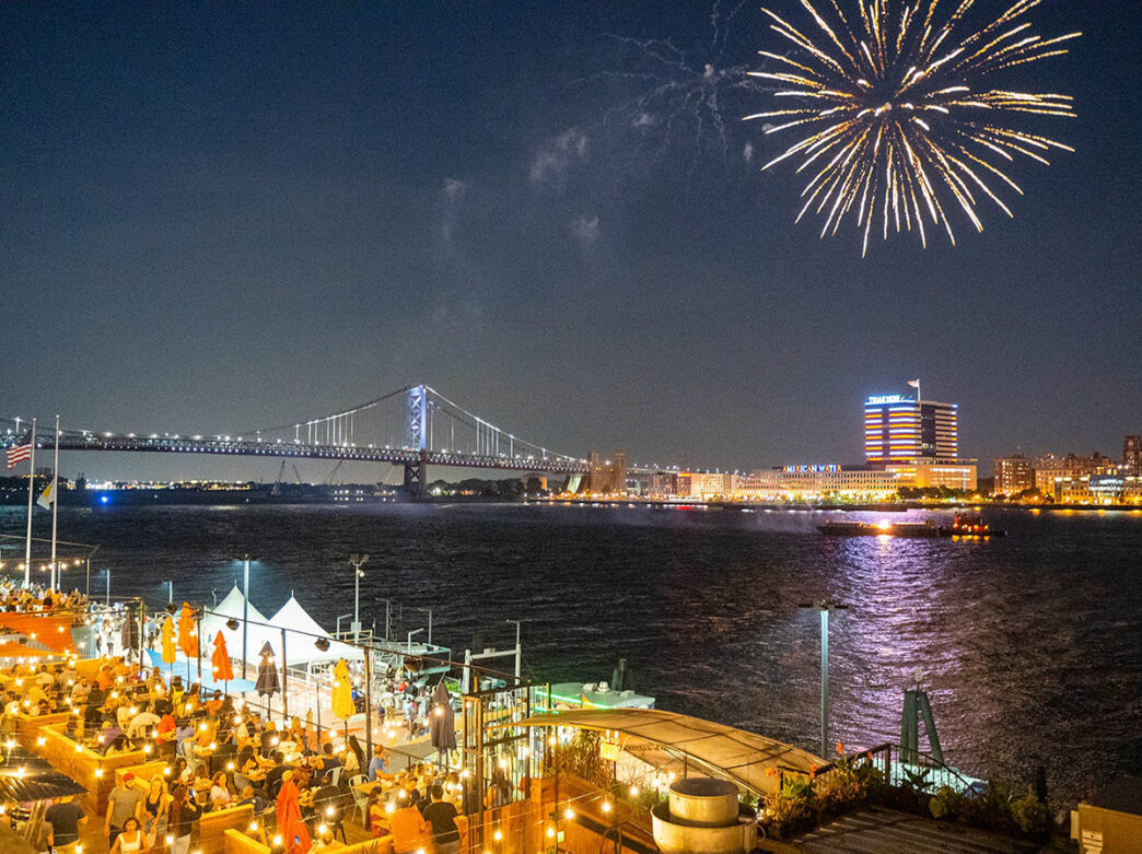 Guests are seated at Liberty Point's outdoor dining space along the Delaware River as a golden firework sparkles over the river with the Benjamin Franklin Bridge in the background.