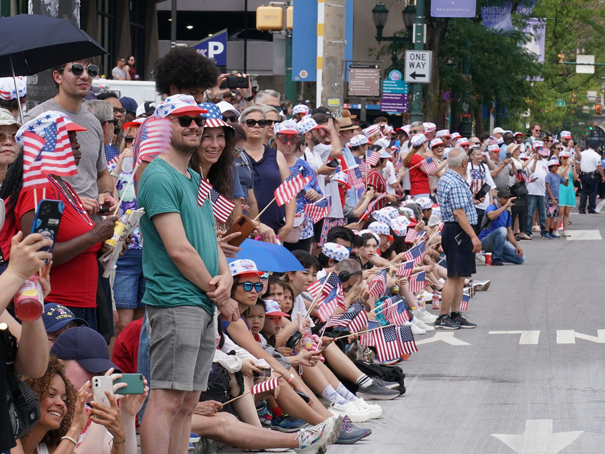 A festive crowd lines the street in Philadelphia for the Fourth of July parade. Many wearing red, white and blue, and waving mini American flags.