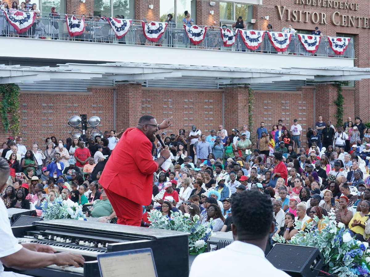 A singer in a bright red suit sings on stage in front of a large crowd outside the Independence Visitor Center, decorated with patriotic bunting.