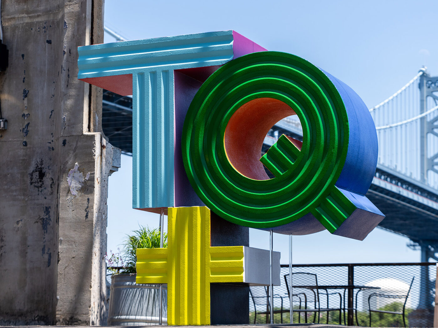 A multi-colored sculpture that says "TQ+" stands outdoors. The Delaware River and Benjamin Franklin Bridge are visible in the background.