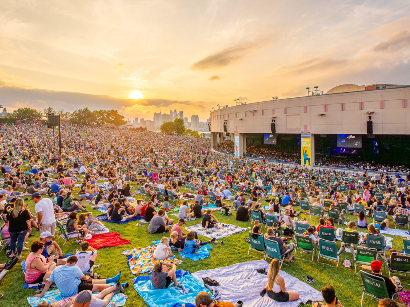 Crowds of people sitting on blankets and lawn chairs fill a large grassy hillside facing the Freedom Mortgage Pavilion during a concert. The Philadelphia skyline and setting sun is seen in the distance.