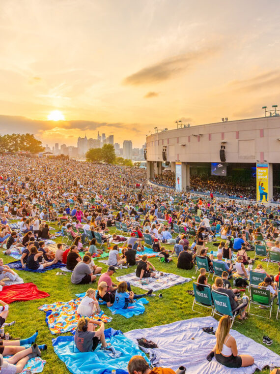 Crowds of people sitting on blankets and lawn chairs fill a large grassy hillside facing the Freedom Mortgage Pavilion during a concert. The Philadelphia skyline and setting sun is seen in the distance.