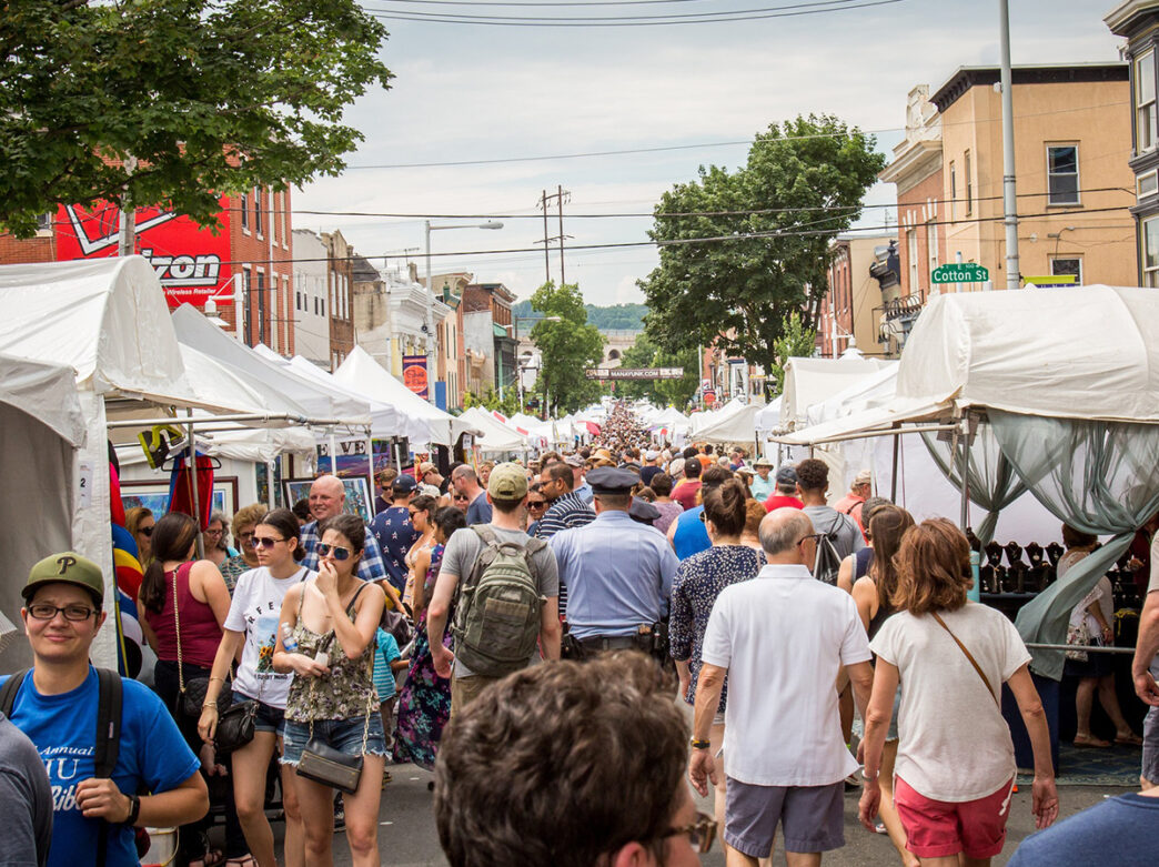 A crowd of people walks through rows of white vendor tents at the Manayunk Arts Festival, browsing artwork and handmade goods.