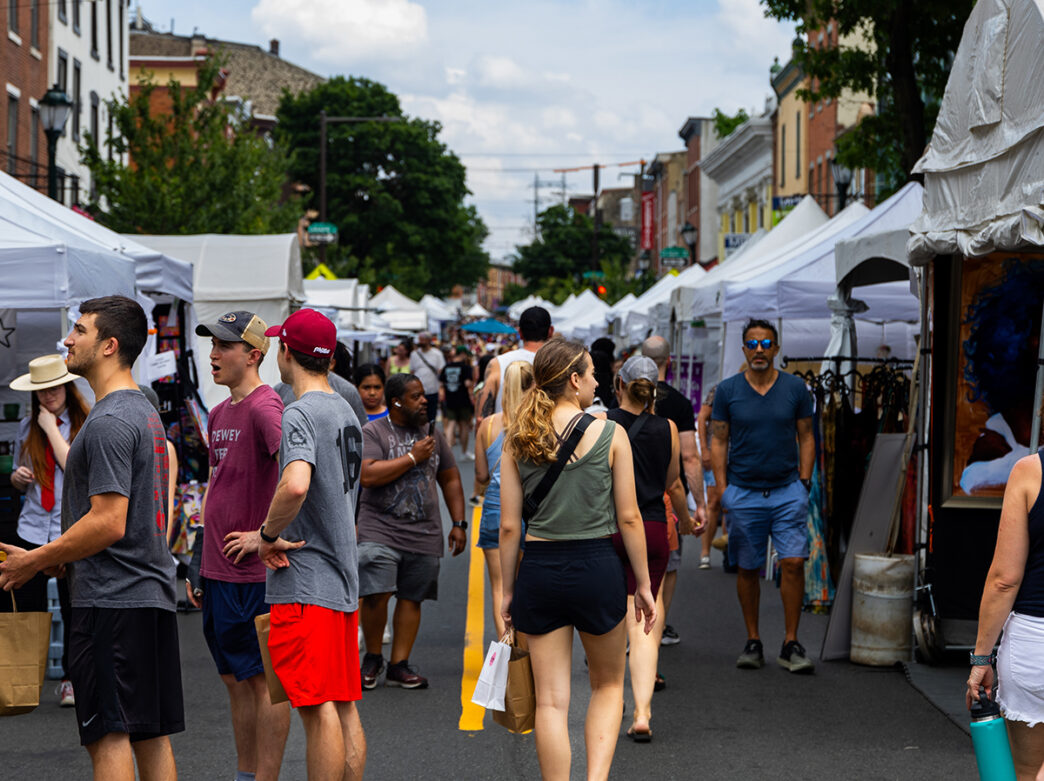 Crowds browse art and crafts at the Manayunk Arts Festival, walking between rows of white vendor tents lining Main Street.