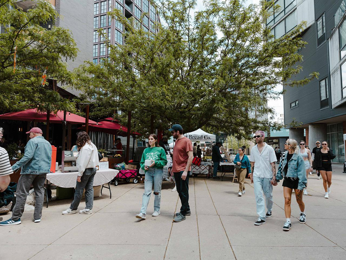 People stroll past vendor tables at an outdoor market in the Piazza in Northern Liberties. Tall apartment buildings surround the plaza.