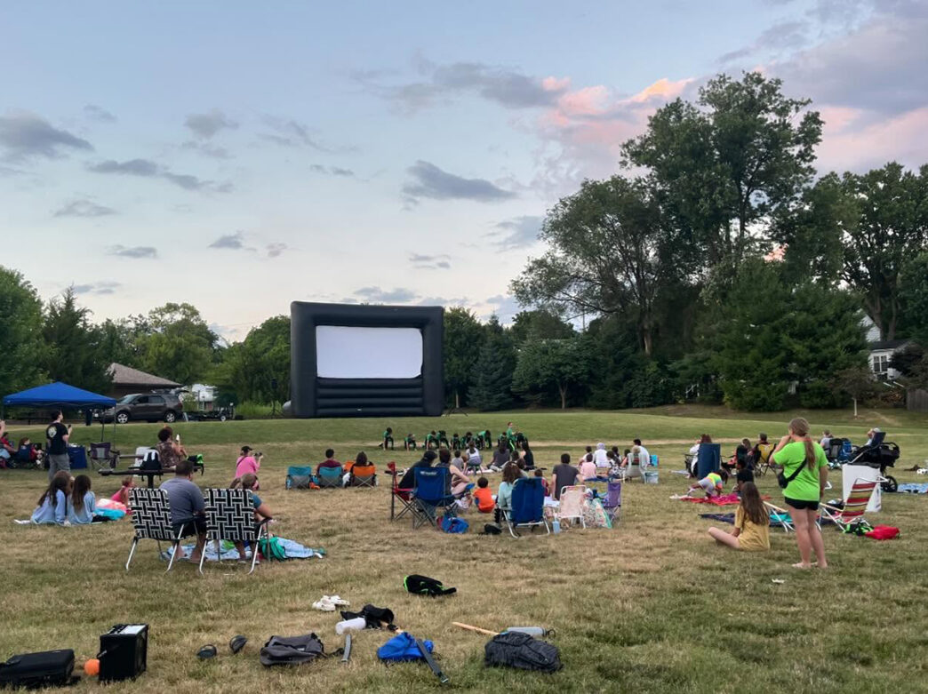Families lounge on blankets and camp chairs in a grassy field, waiting for an outdoor movie to start on a large inflatable screen.