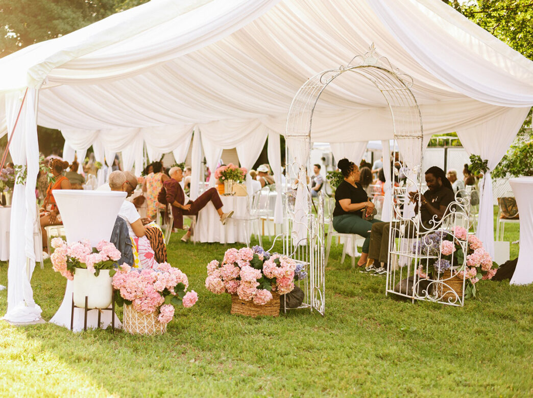 People relax under a white draped tent adorned with pink flowers at the Navy Yard Wine Garden, enjoying drinks and conversation.