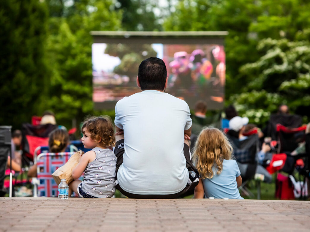 A man and two young children sit on the ground facing an outdoor movie screen surrounded by a crowd of people in folding chairs.