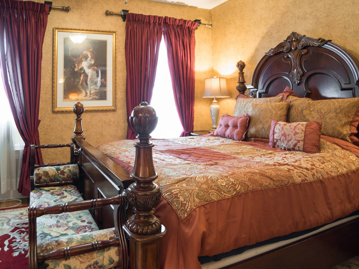 A richly decorate guest room features an ornate wooden bed with golden and red bedding and matching red curtains.